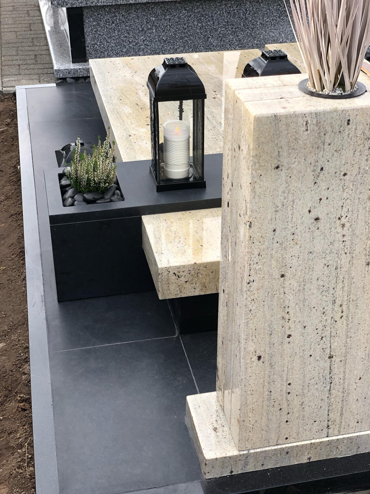 Jasny, nowoczesny pomnik z granitu z czarnymi akcentami, lampionem i roślinami. Minimalistyczny design, eleganckie wykończenie.