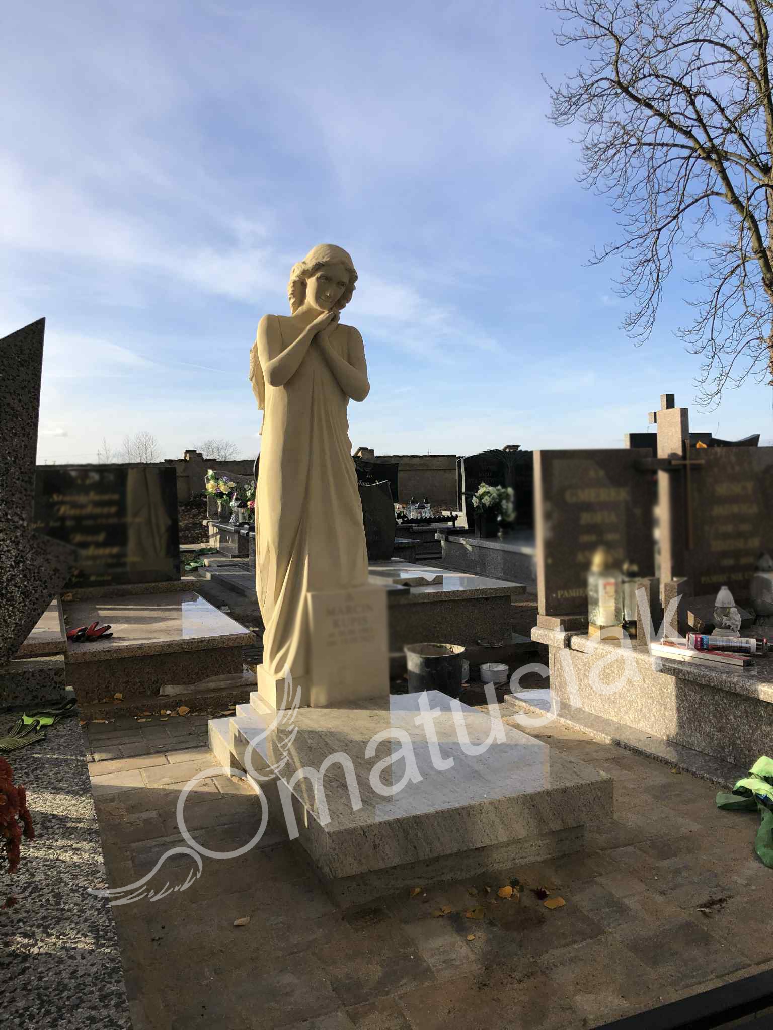 Jasny nagrobek z rzeźbą anioła na cmentarzu, w tle inne groby i drzewo bez liści. Nagrobek wykonany z jasnego kamienia.