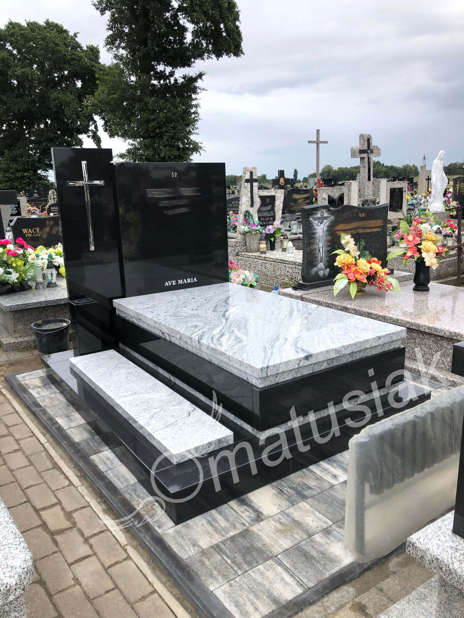 Czarny, nowoczesny pomnik z jasnoszarym blatem i krzyżem, na cmentarzu. Widoczne inne groby i krzyże w tle. Kamienna kostka brukowa wokół.