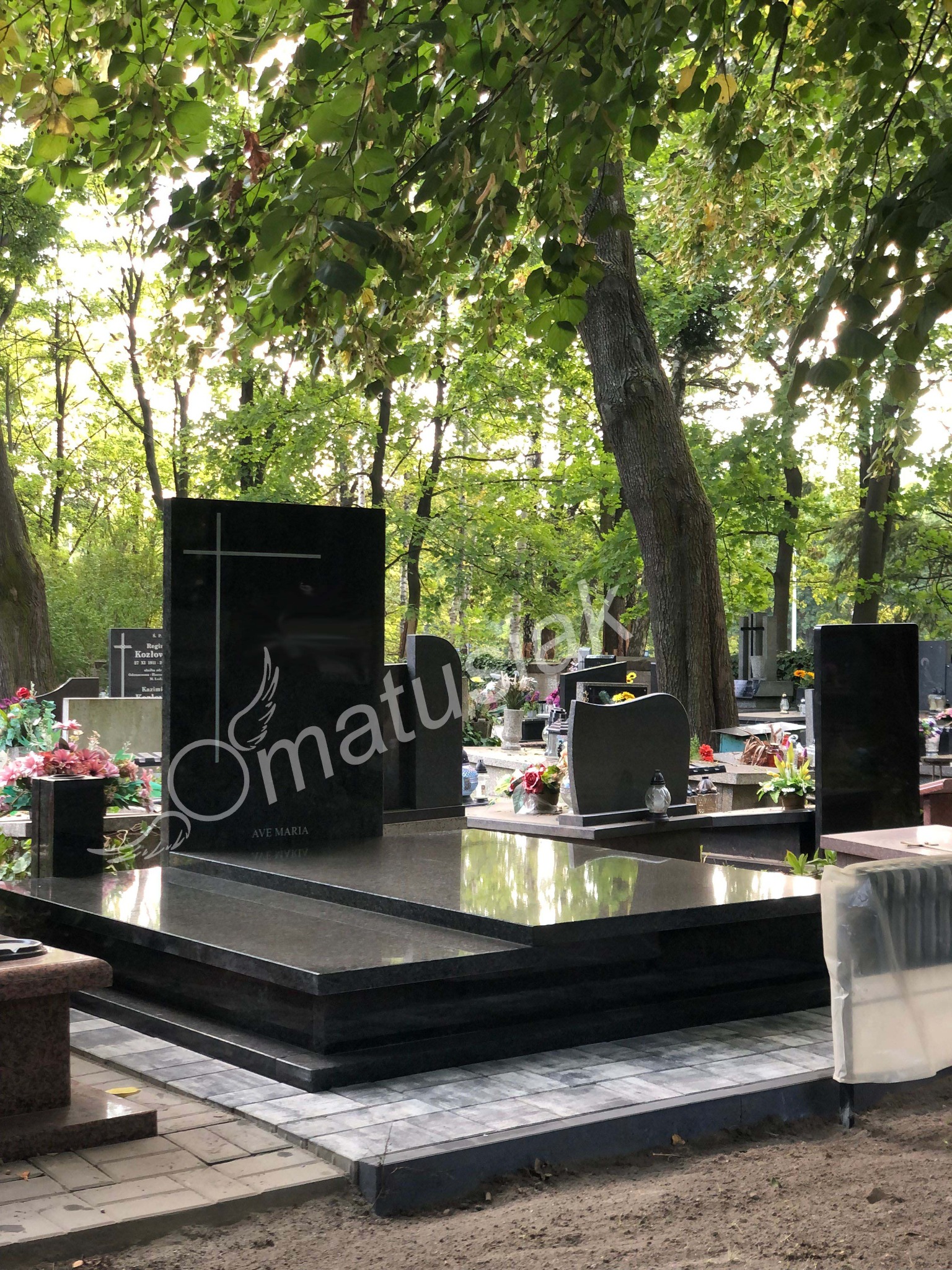 Czarny, polerowany pomnik z krzyżem i napisem 'Ave Maria' na tle cmentarza z drzewami. Nagrobek odbija światło, tworząc lustrzany efekt.