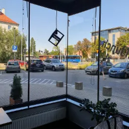 Widok z wnętrza budynku przez duże, czyste okno na parking z samochodami, drzewami i budynkami w tle. Na szybie widoczne naklejki i odbicia. Wnętrze lokalu zdobi nowoczesna lampa wisząca.