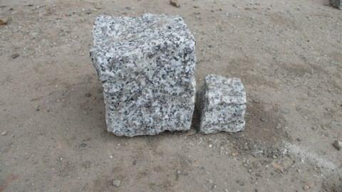 Dwie kostki granitowe o nieregularnych kształtach na piaszczystym podłożu. Większa kostka stoi obok mniejszej, tworząc kontrast wielkości.