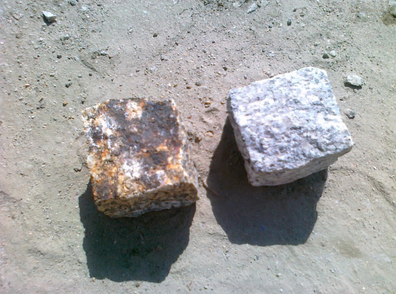 Dwie kostki brukowe, jedna szara, druga brązowa, leżą na piasku. Widoczne nierówności i faktura materiału. Oświetlenie słoneczne podkreśla detale.