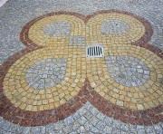 Nawierzchnia z kostki granitowej ułożona w dekoracyjny wzór czterolistnej koniczyny wokół kratki kanalizacyjnej. Użyto kostki w odcieniach szarości, beżu i brązu.