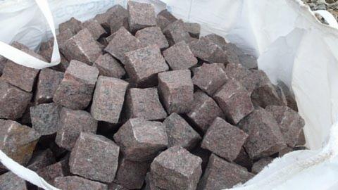 Czerwona kostka granitowa w worku typu big bag. Kostka o nieregularnych krawędziach, widoczne ziarna granitu. Jasne tło worka kontrastuje z kolorem kamienia.