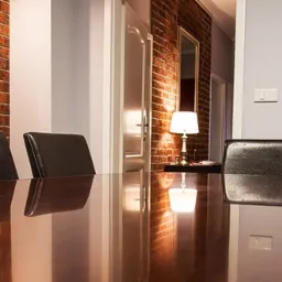 Sala konferencyjna z dużym, błyszczącym stołem, skórzanymi krzesłami i ścianą z czerwonej cegły w tle, oświetlona lampą stołową.