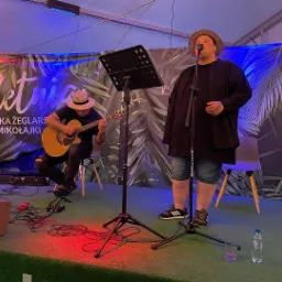 Duet muzyczny na scenie: piosenkarz w słomkowym kapeluszu śpiewa do mikrofonu, gitarzysta gra na akustycznej gitarze, w tle baner z napisem 'Na Letnia Wioska Żeglarska Mikołajki'.