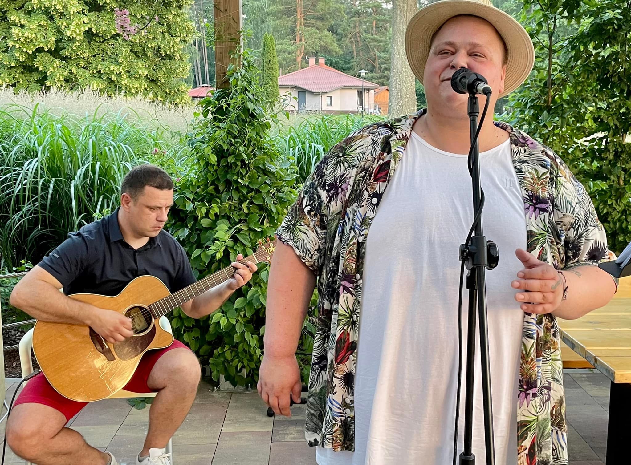 Wokalista w kwiecistej koszuli i słomkowym kapeluszu śpiewa do mikrofonu na tle gitarzysty grającego na gitarze akustycznej podczas występu plenerowego.