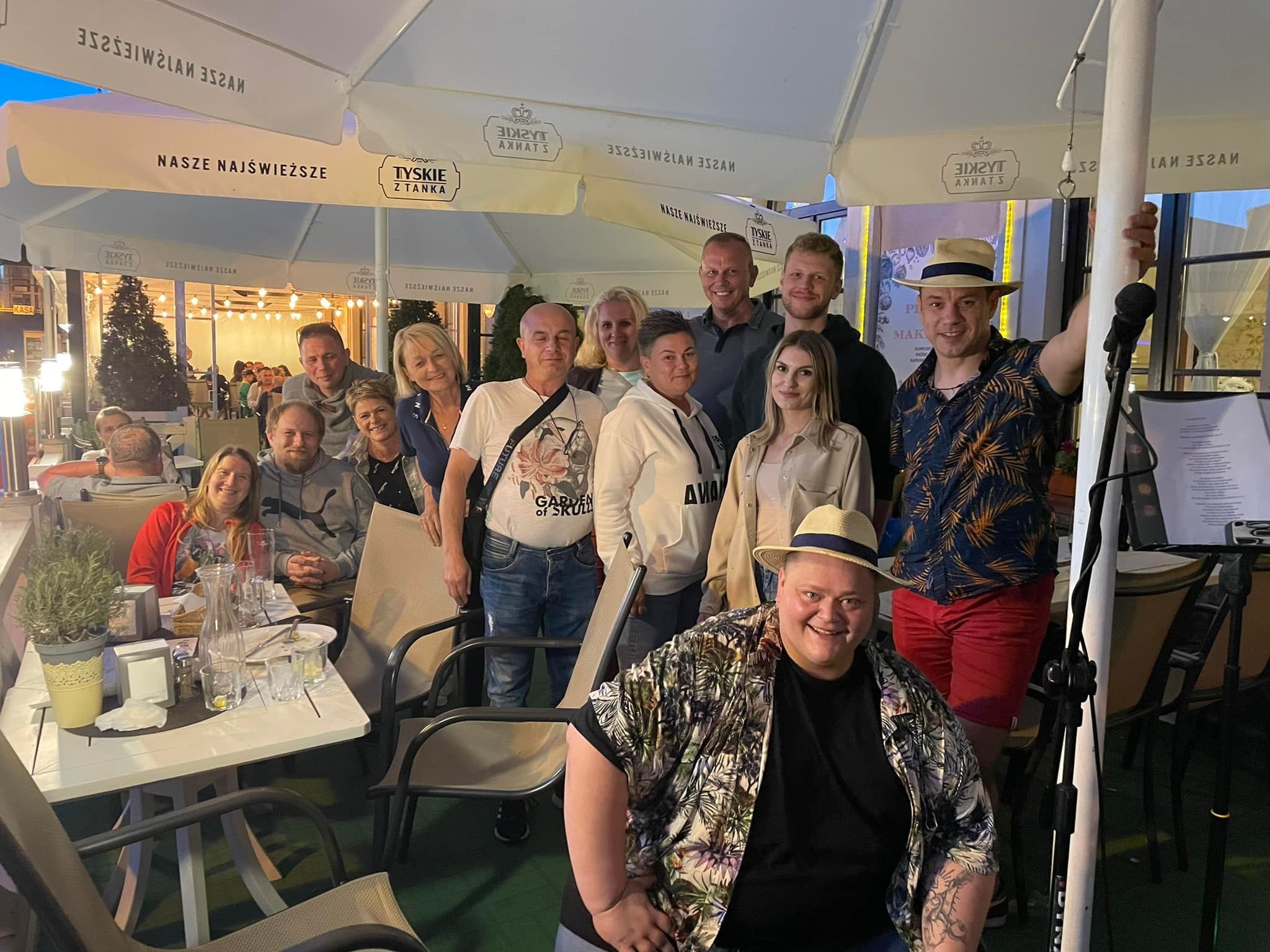 Grupa osób pozuje do zdjęcia na zewnątrz, pod parasolami reklamowymi Tyskie, widoczny mikrofon na statywie, sugerujący występ muzyczny.