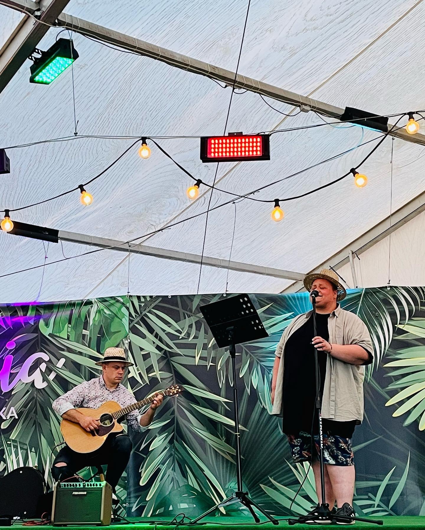Duet muzyczny na scenie pod namiotem: wokalista w słomkowym kapeluszu śpiewa do mikrofonu, gitarzysta gra na akustycznej gitarze, w tle tropikalna dekoracja i oświetlenie sceniczne.