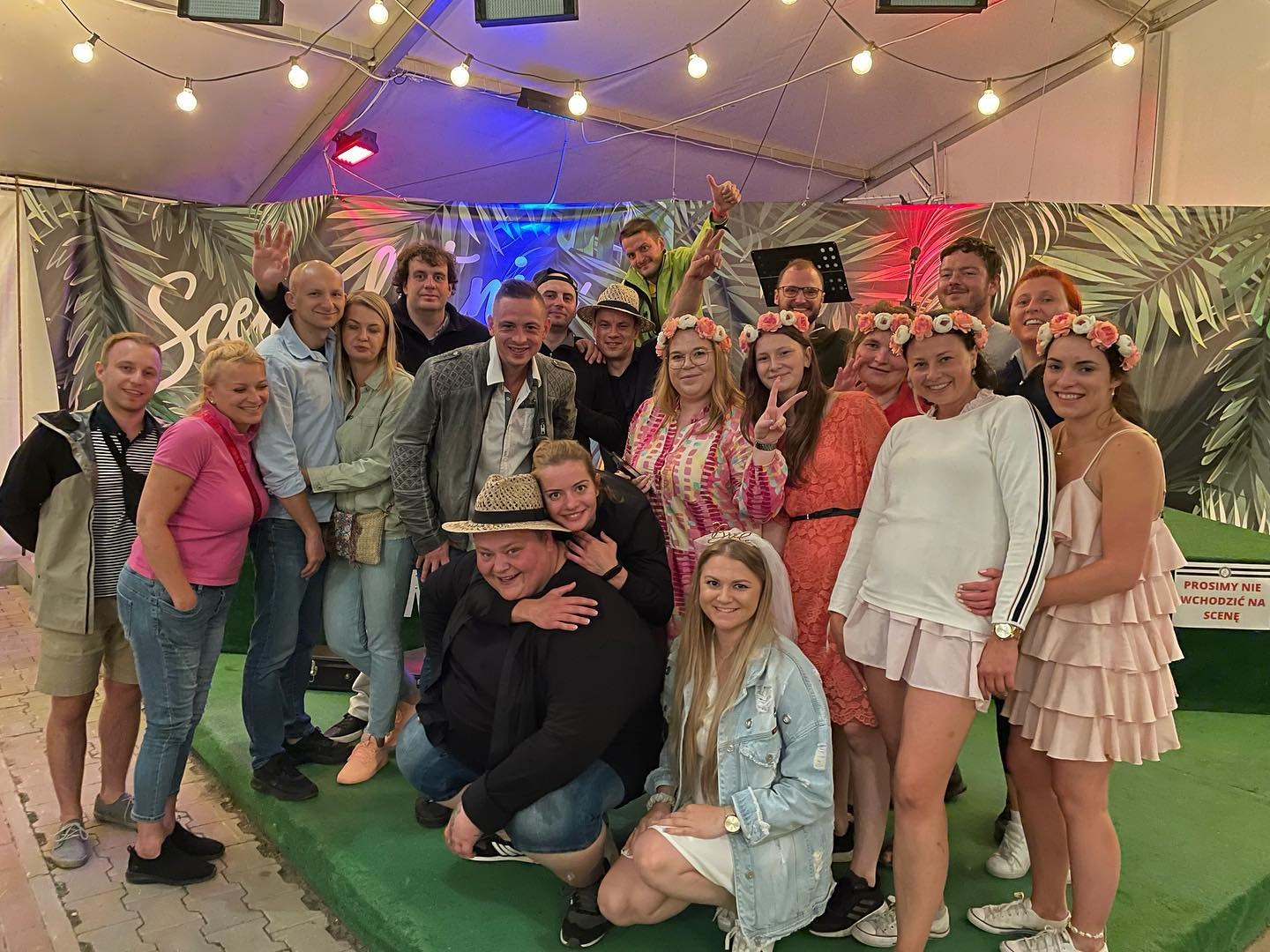 Grupa uśmiechniętych osób pozuje na scenie z dekoracją z liści, część osób ma wianki na głowach, a jedna osoba ma welon na głowie. W tle widoczny napis 'Prosimy nie wchodzić na scenę'.