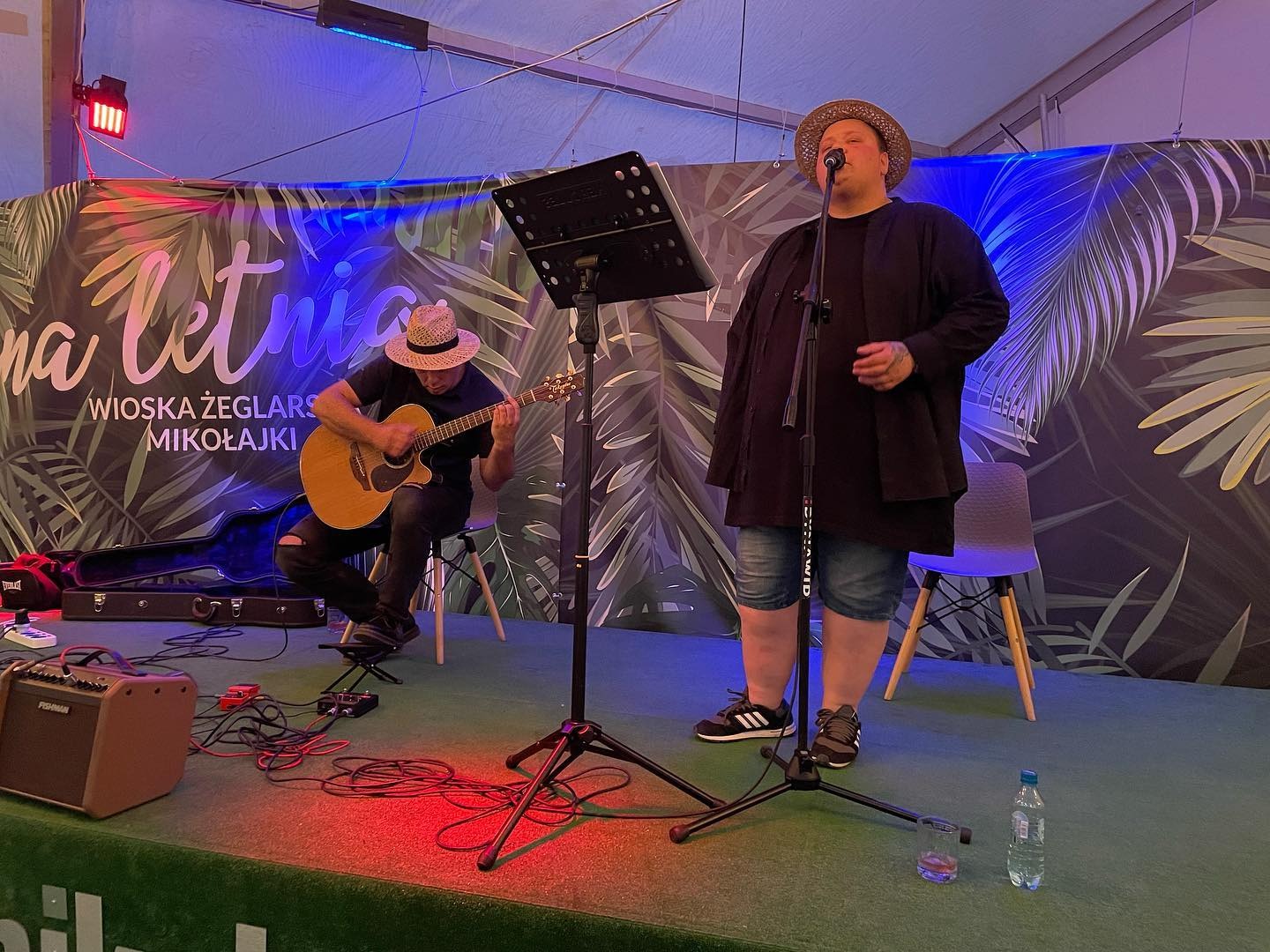 Duet muzyczny na scenie: piosenkarz w słomkowym kapeluszu śpiewa do mikrofonu, gitarzysta gra na akustycznej gitarze, w tle baner z napisem 'Na Letnia Wioska Żeglarska Mikołajki'.