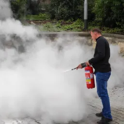 Mężczyzna w jeansach i czarnym swetrze demonstruje użycie czerwonej gaśnicy proszkowej na zewnątrz, tworząc gęsty obłok białego dymu na tle kamiennego muru i zieleni, z dwiema innymi gaśnicami...