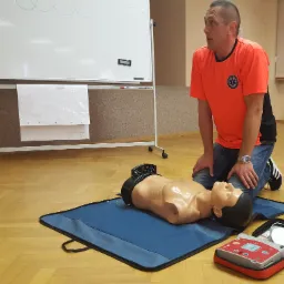 Instruktor na kolanach obok fantoma do resuscytacji i defibrylatora AED podczas demonstracji. Na koszulce logo ratownictwa medycznego.