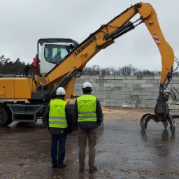 Żółta maszyna Liebherr z chwytakiem hydraulicznym, dwóch inspektorów UDT w kamizelkach odblaskowych i kaskach obserwuje pracę operatora w kabinie, w tle bloki betonowe