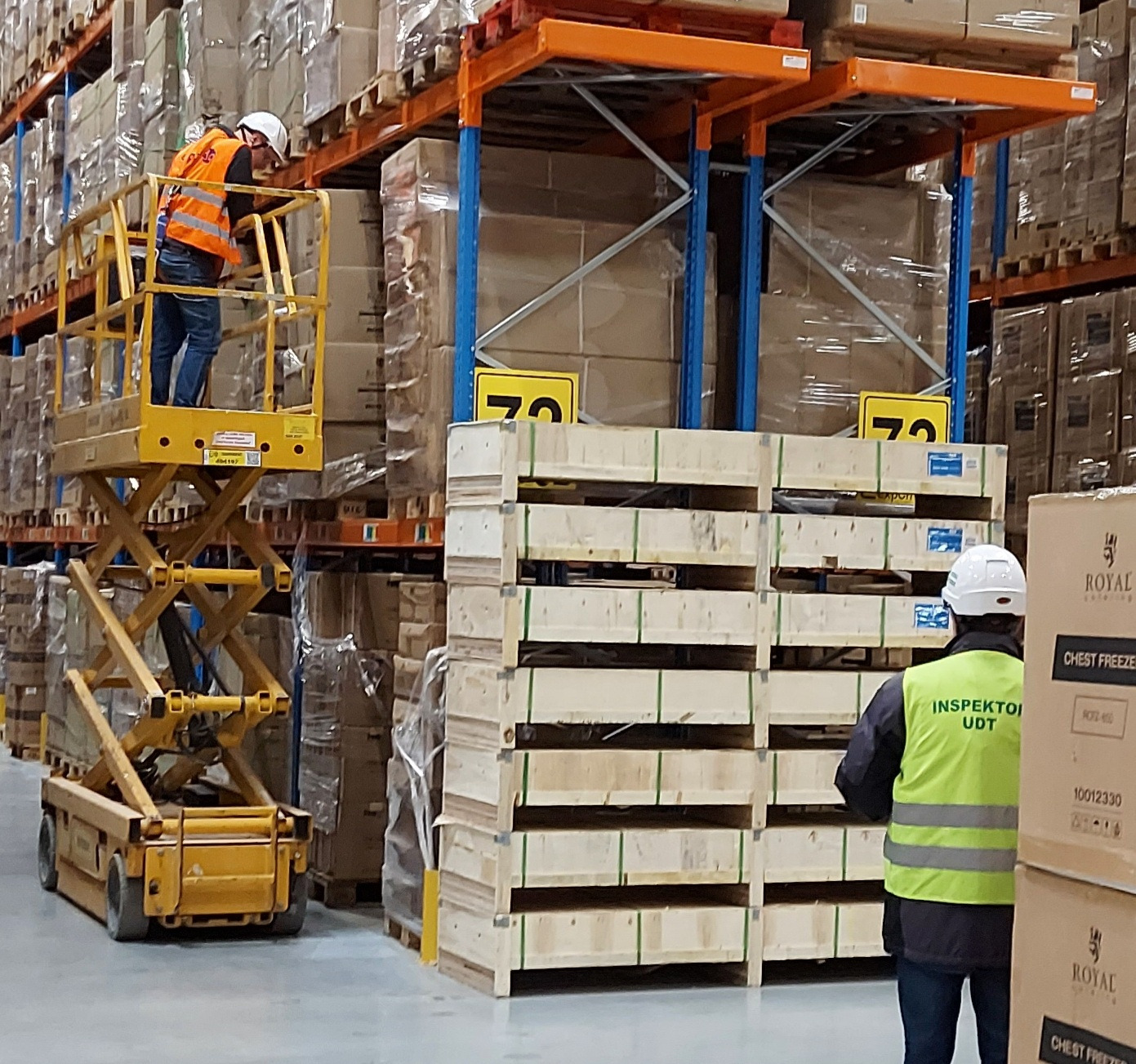 Inspektor UDT w kamizelce i kasku kontroluje pracę pracownika na podnośniku nożycowym w magazynie wysokiego składowania z paletami i regałami.