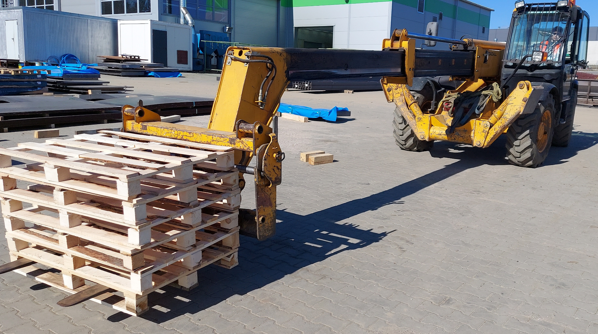 Żółty wózek widłowy teleskopowy podnosi stos jasnych, drewnianych palet na zewnątrz, na tle magazynu przemysłowego.