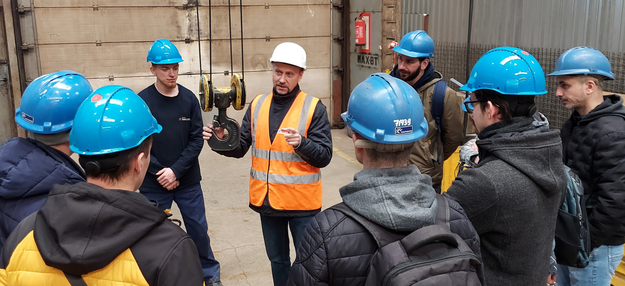Instruktor w pomarańczowej kamizelce prezentuje hak dźwigowy grupie osób w niebieskich i białych kaskach na tle hali produkcyjnej.