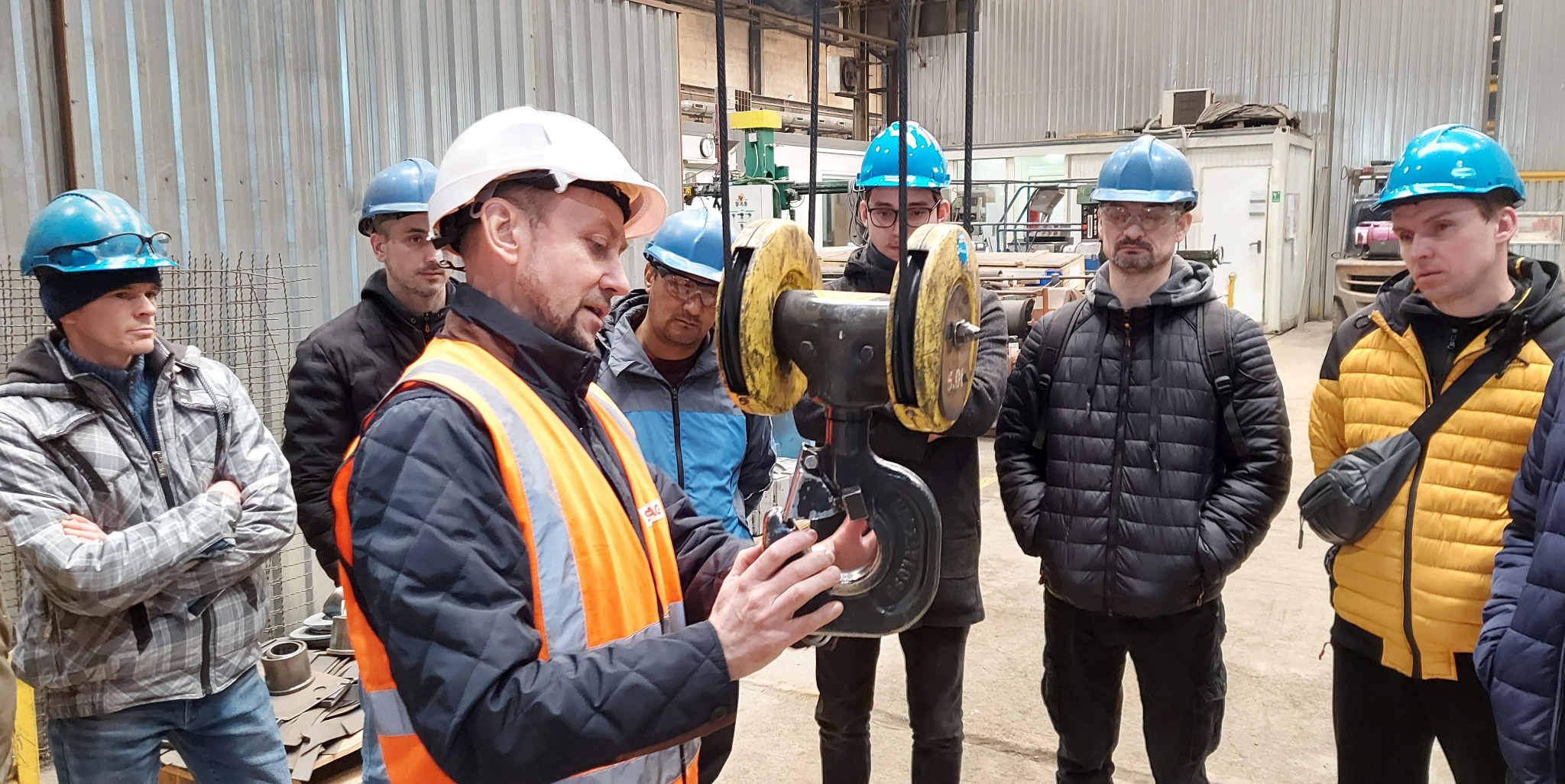 Instruktor w pomarańczowej kamizelce omawia z grupą kursantów w hełmach budowę i działanie zawiesia hakowego w hali produkcyjnej w Zielonej Górze.