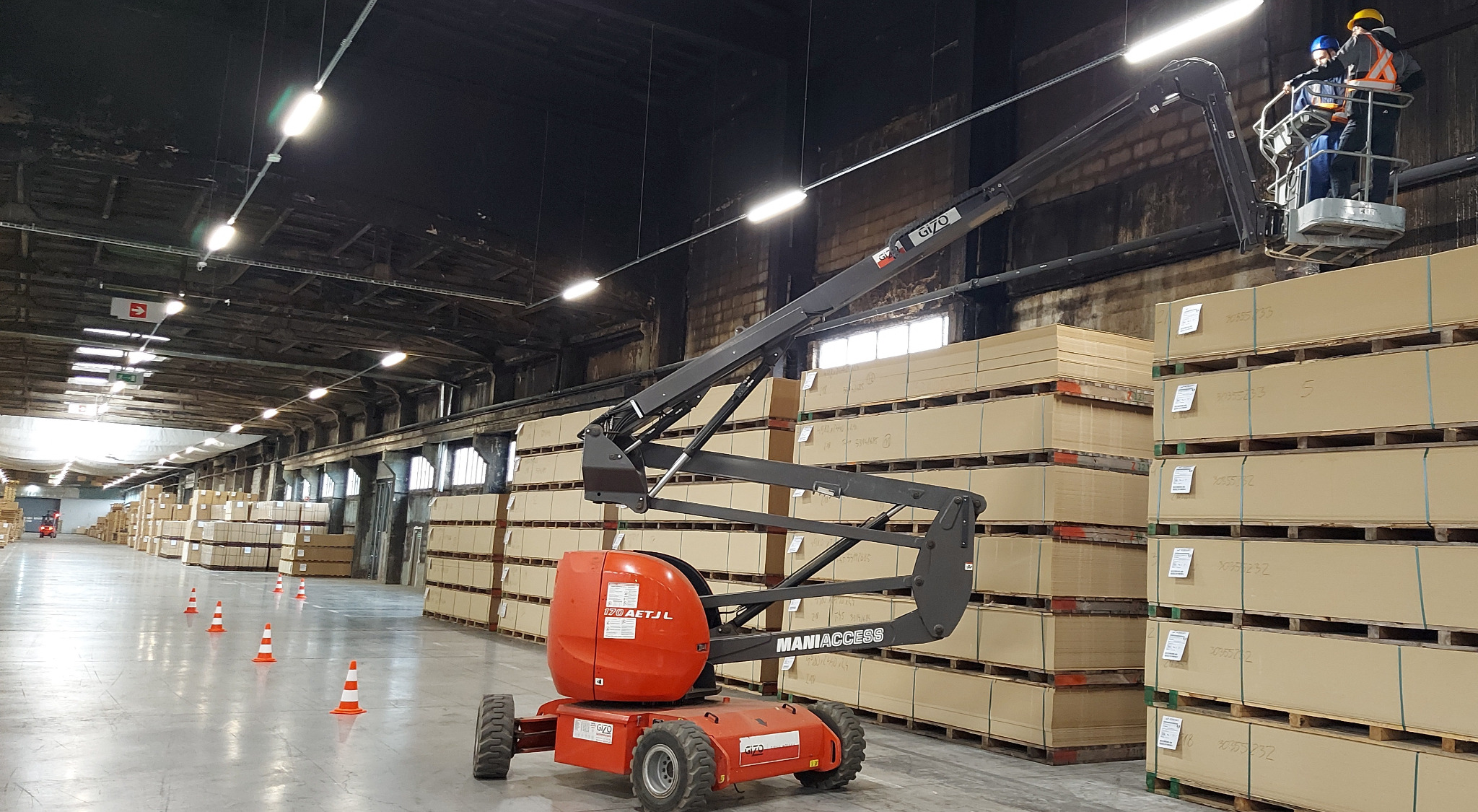 Wnętrze magazynu z podnośnikiem koszowym Maniaccess 170 AETJL, na którym znajdują się dwaj pracownicy w kaskach i kamizelkach odblaskowych, wykonujący prace na wysokości wśród palet z towarem...