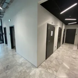 Korytarz biurowy z szarymi drzwiami, marmurową podłogą i czarnym sufitem z podłużnymi lampami LED.