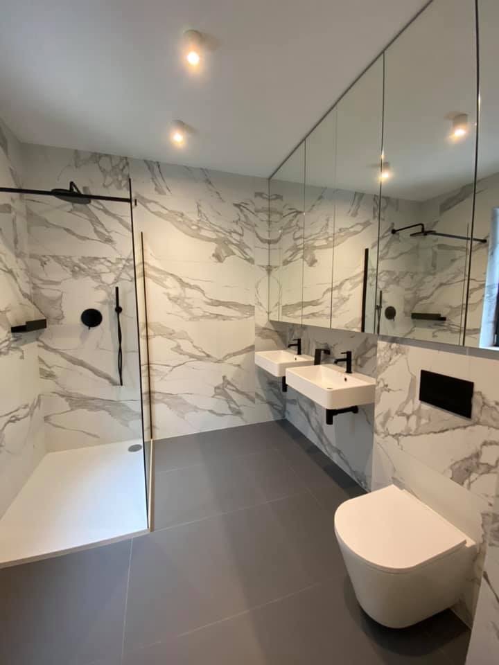 Nowoczesna łazienka z marmurowymi płytkami ściennymi, szarą podłogą, kabiną prysznicową typu walk-in z czarną armaturą, podwójną umywalką nablatową i wiszącą toaletą.