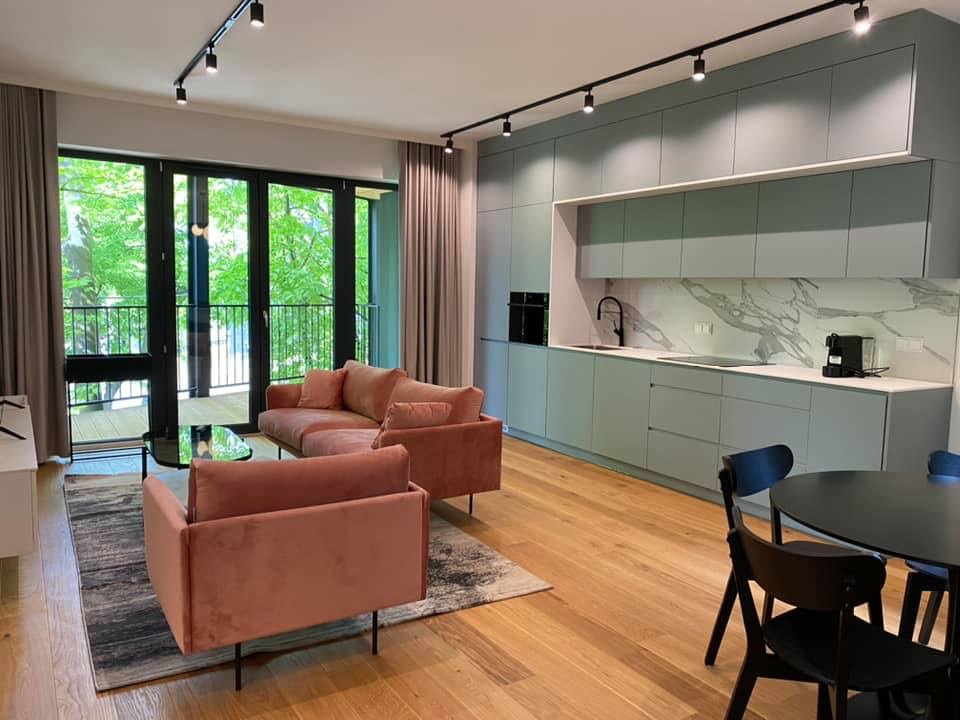 Nowoczesny salon z otwartą kuchnią: pastelowe szafki kuchenne z marmurowym backsplash, różowa sofa i fotel na tle dużego okna balkonowego.