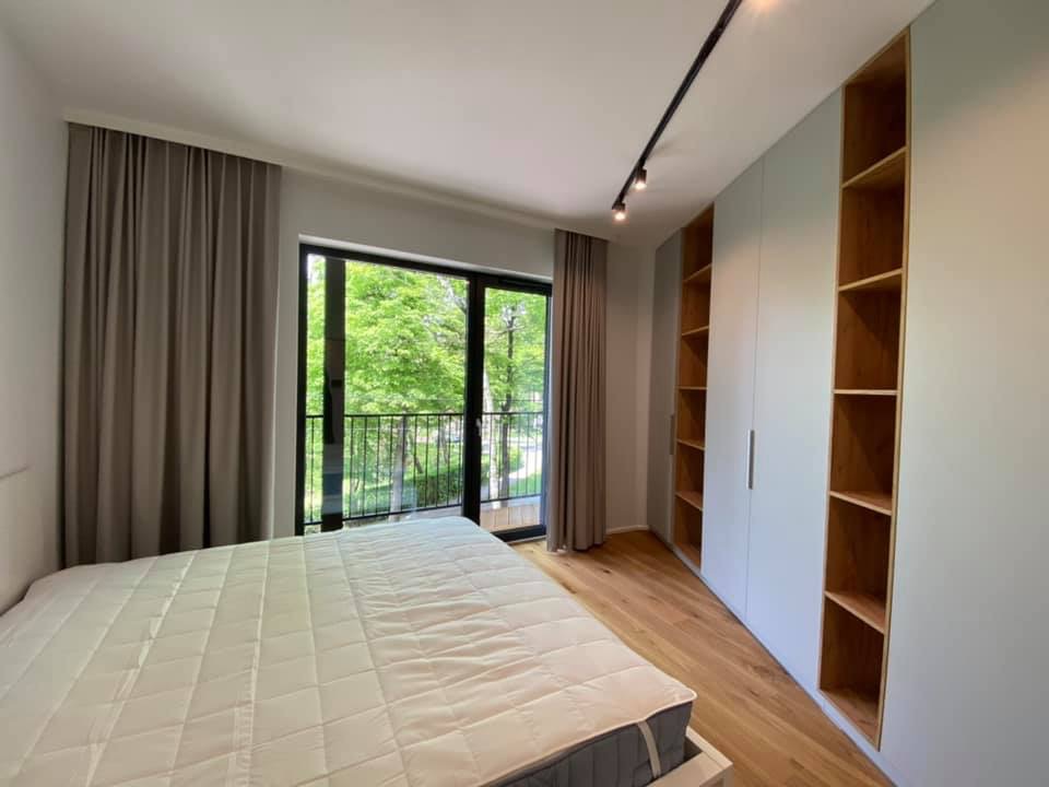 Jasna sypialnia z dużym oknem balkonowym z widokiem na zieleń, drewnianą podłogą i zabudowaną szafą z otwartymi półkami.