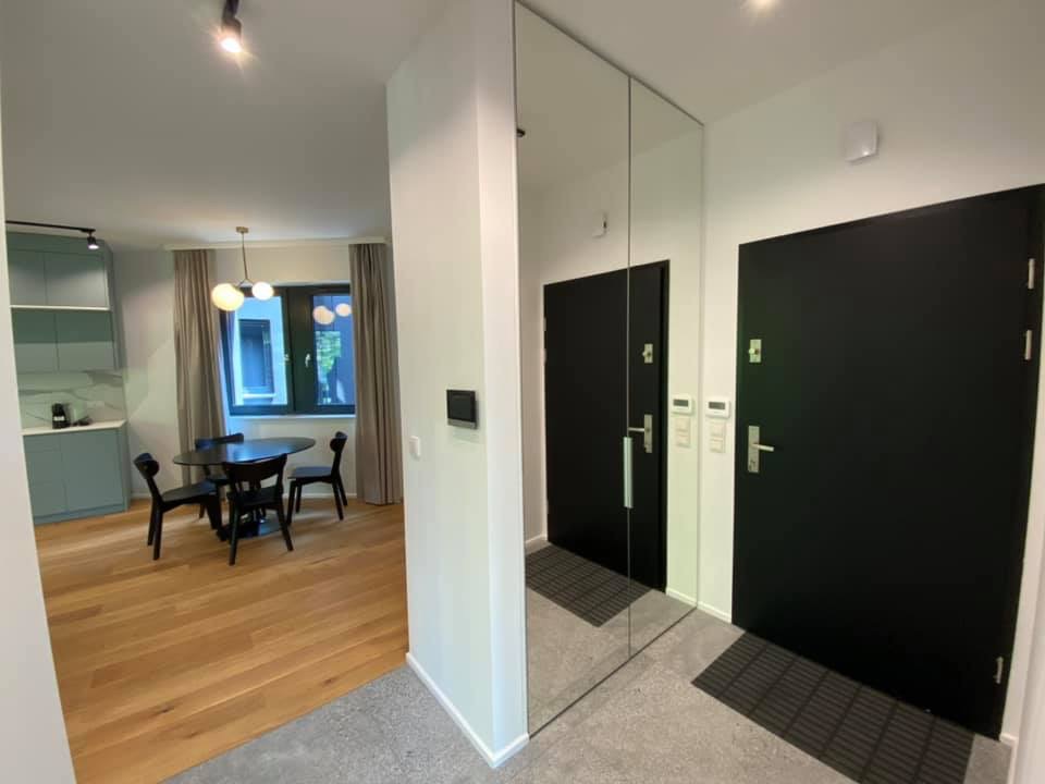 Nowoczesne wnętrze mieszkania z parkietem, czarnym stołem i krzesłami, oraz lustrzaną ścianą obok czarnych drzwi wejściowych.