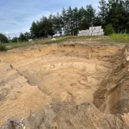 Szeroki wykop pod fundamenty w Wągrowcu, z widocznymi śladami po pracy maszyn i stosem bloczków betonowych w tle.