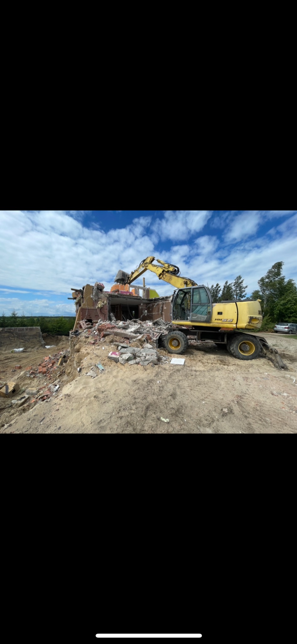 Żółta koparka kołowa z wysięgnikiem kruszy resztki ceglanego budynku na piaszczystym terenie pod błękitnym niebem z chmurami.