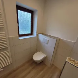 Nowoczesna łazienka po remoncie: podwieszana toaleta z przyciskiem spłukiwania, drewnopodobna podłoga, grzejnik drabinkowy, okno z teksturowaną szybą i dekoracyjne wykończenie nad zabudową toalety.