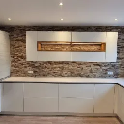 Nowoczesna kuchnia z białymi, minimalistycznymi szafkami, blatem w jasnym odcieniu i dekoracyjną ścianą z imitacji kamienia, podświetlane półki.