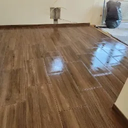 Ułożone, brązowe płytki podłogowe imitujące drewno, z widocznymi refleksami światła, w trakcie prac wykończeniowych, z workiem na śmieci w tle.