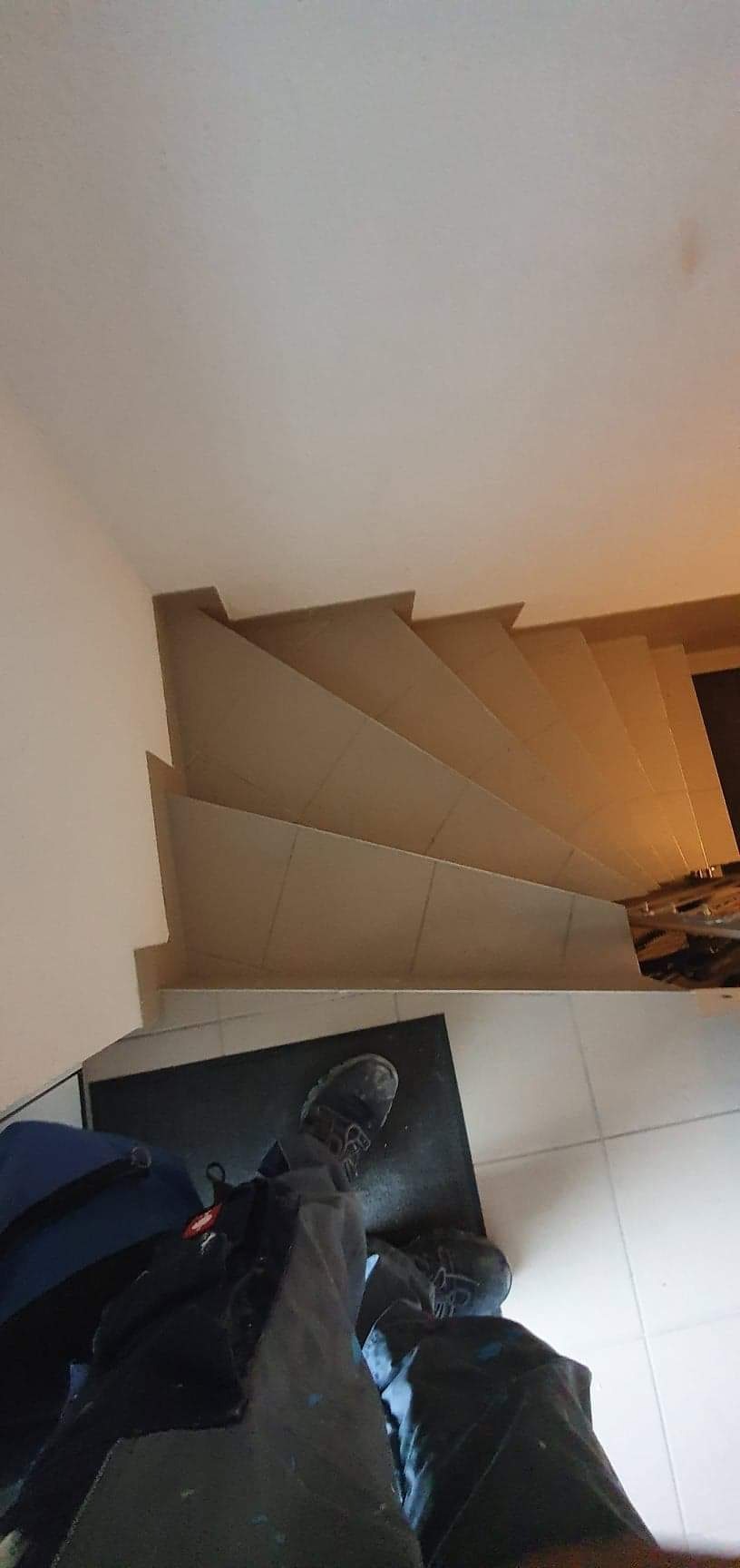 Wyłożone płytkami schody w trakcie remontu, widok z góry na stopy robotnika, na podłodze widoczny worek i narzędzia.