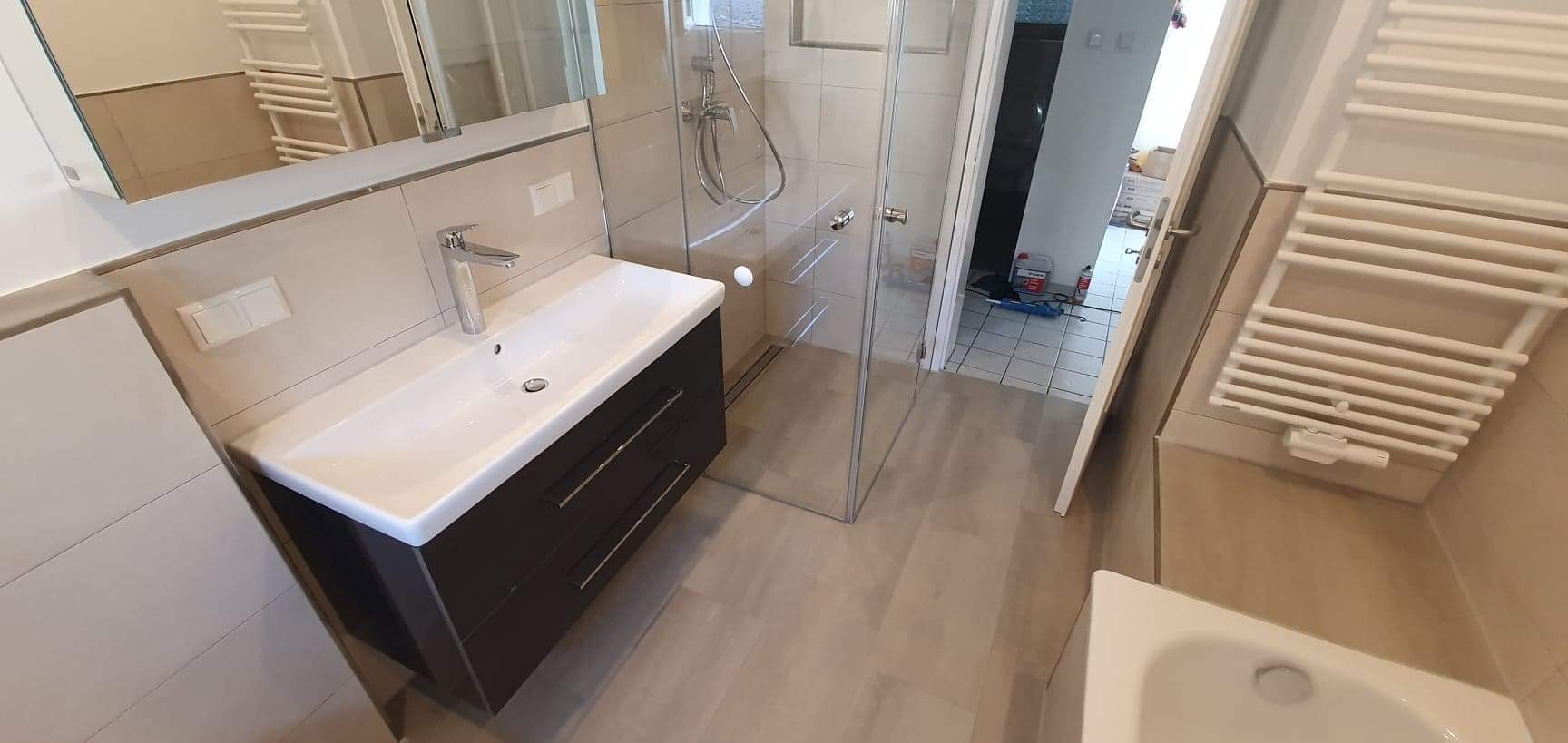 Wykończona łazienka z kabiną prysznicową typu walk-in, umywalką z szafką w kolorze ciemnego brązu, grzejnikiem drabinkowym i płytkami w odcieniach beżu i szarości.