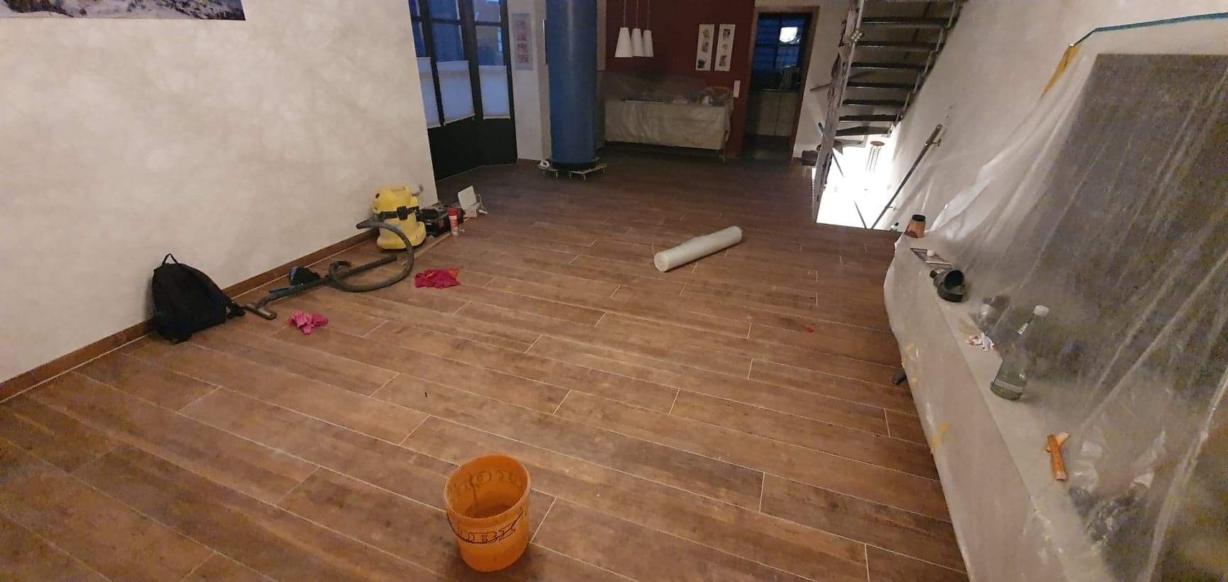 Ułożona podłoga z płytek imitujących drewno w trakcie prac wykończeniowych, widoczne narzędzia, wiadro, odkurzacz i folia ochronna zabezpieczająca elementy wystroju.