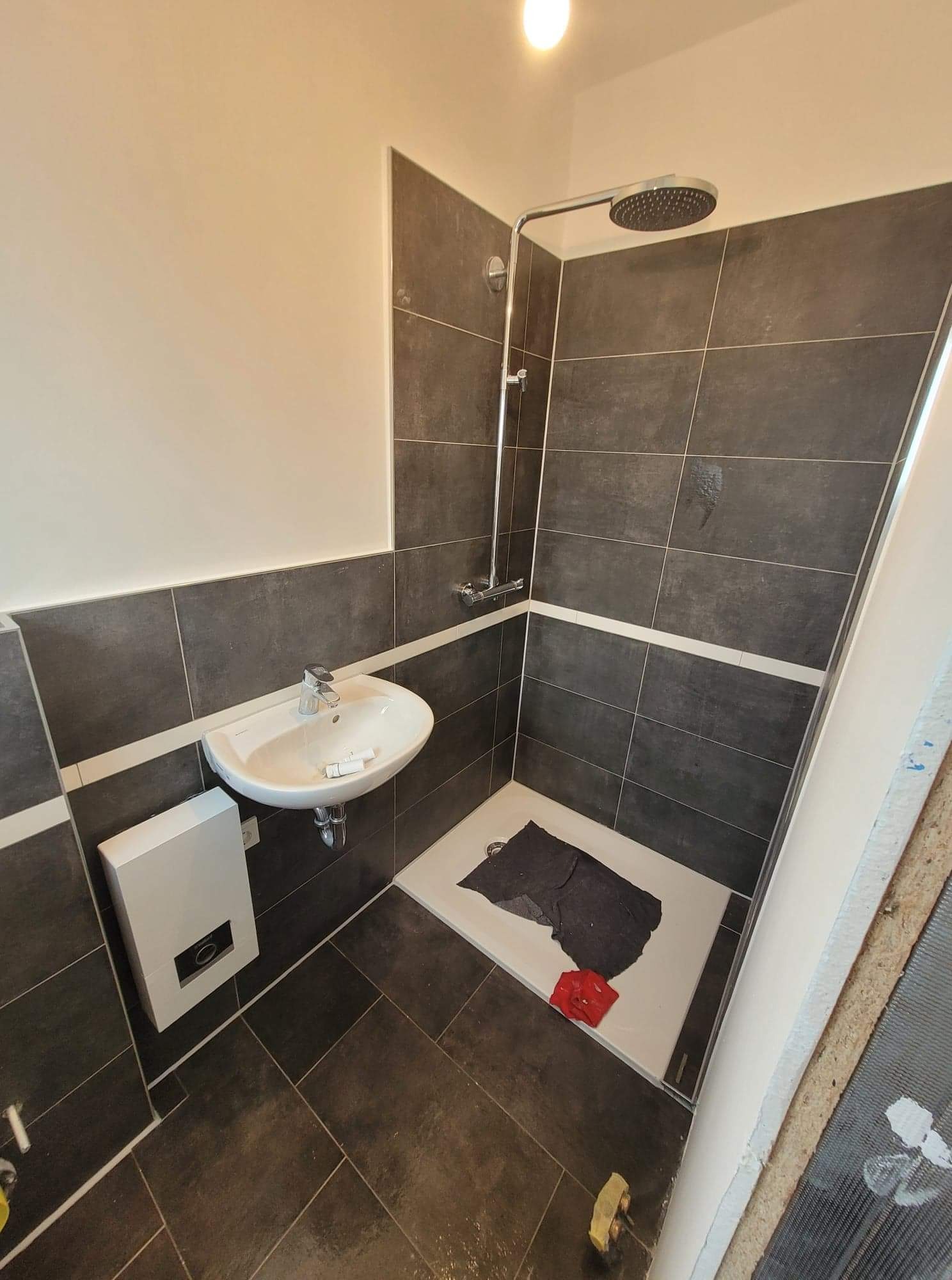 Nowoczesna łazienka z ciemnymi płytkami i białymi akcentami, kabina prysznicowa typu walk-in, umywalka nablatowa i podgrzewacz wody na ścianie, widoczne narzędzia i materiały budowlane sugerują...