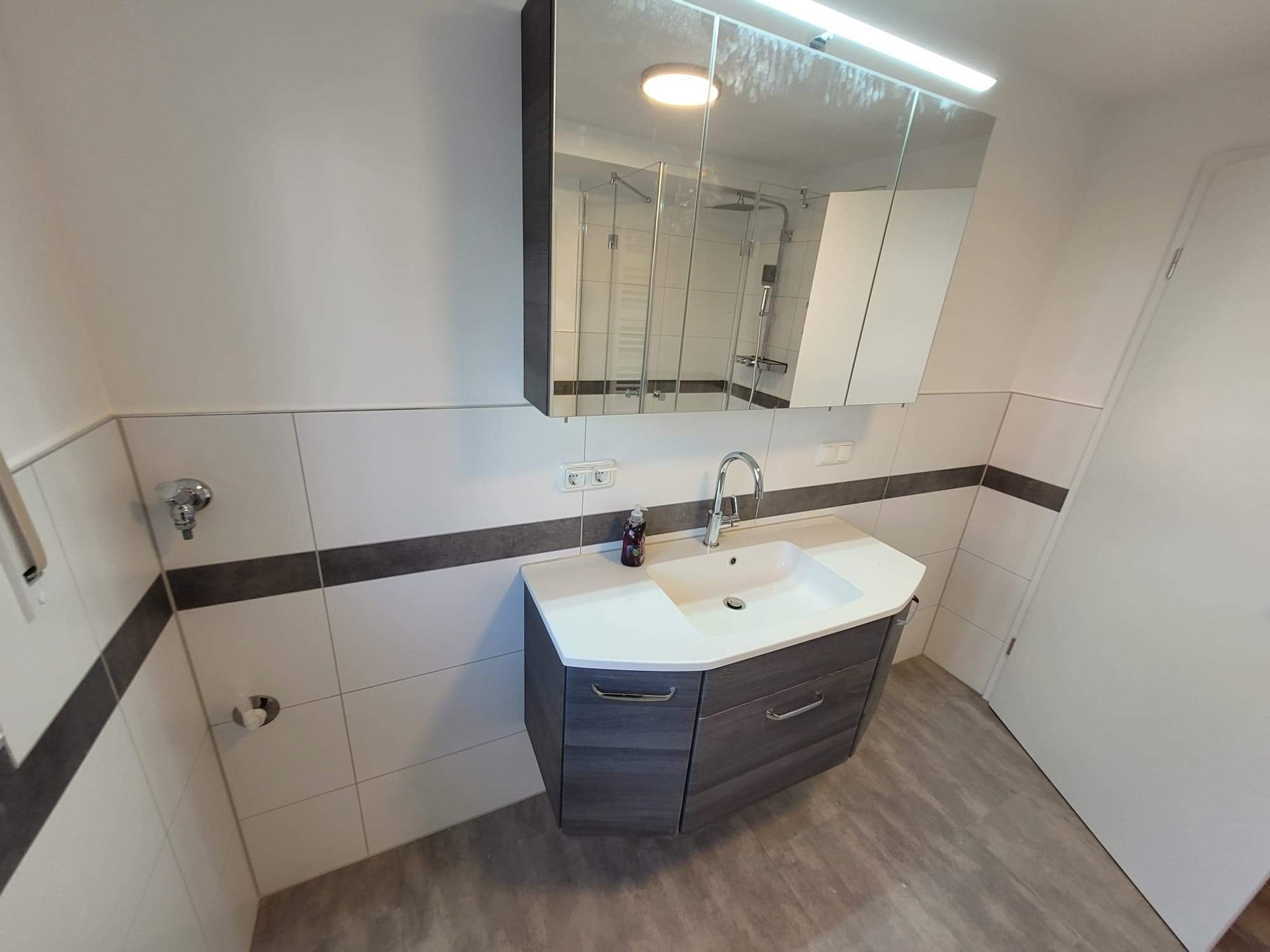 Nowoczesna łazienka z umywalką w szafce z szufladami o fakturze drewna, białą armaturą i lustrem z oświetleniem LED, widoczna butelka mydła w płynie.