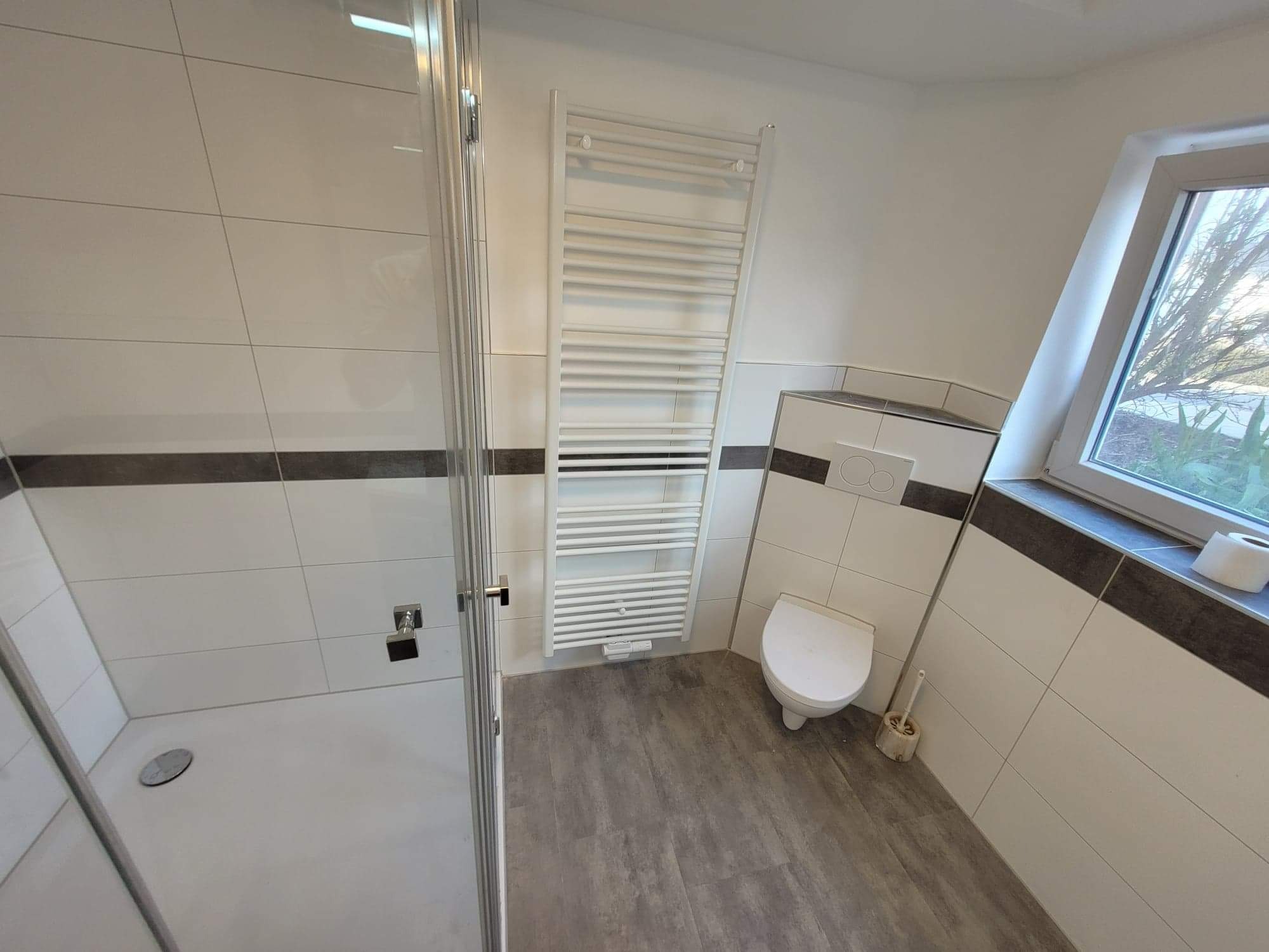 Nowoczesna łazienka po remoncie: kabina prysznicowa ze szklanymi drzwiami, białe płytki z ciemnoszarym pasem, grzejnik drabinkowy, podwieszana toaleta i okno z widokiem na zieleń.