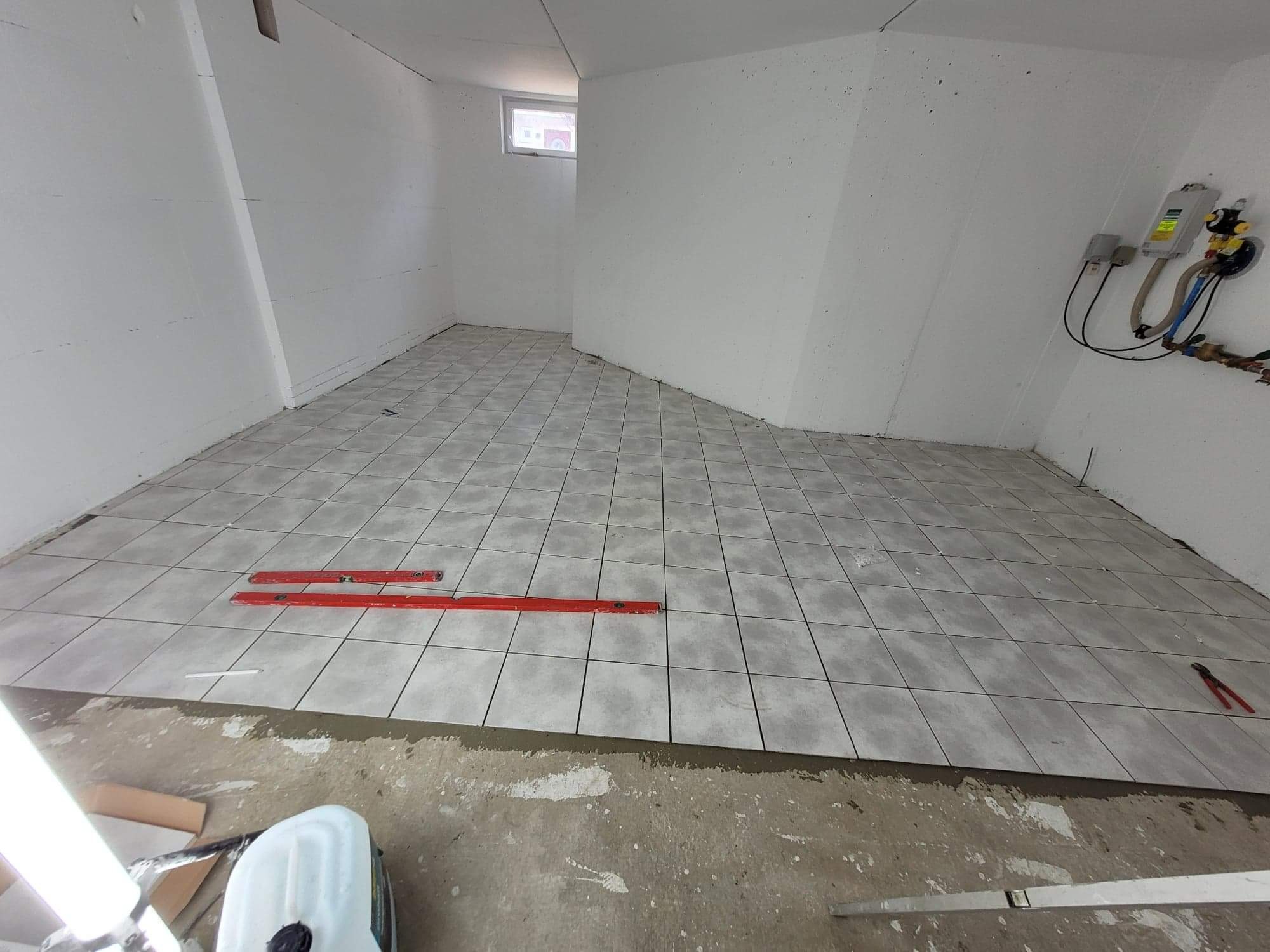 Układanie szarych płytek podłogowych w pomieszczeniu z surowymi, białymi ścianami; widoczne poziomice, pojemnik z klejem i szczypce do cięcia płytek.