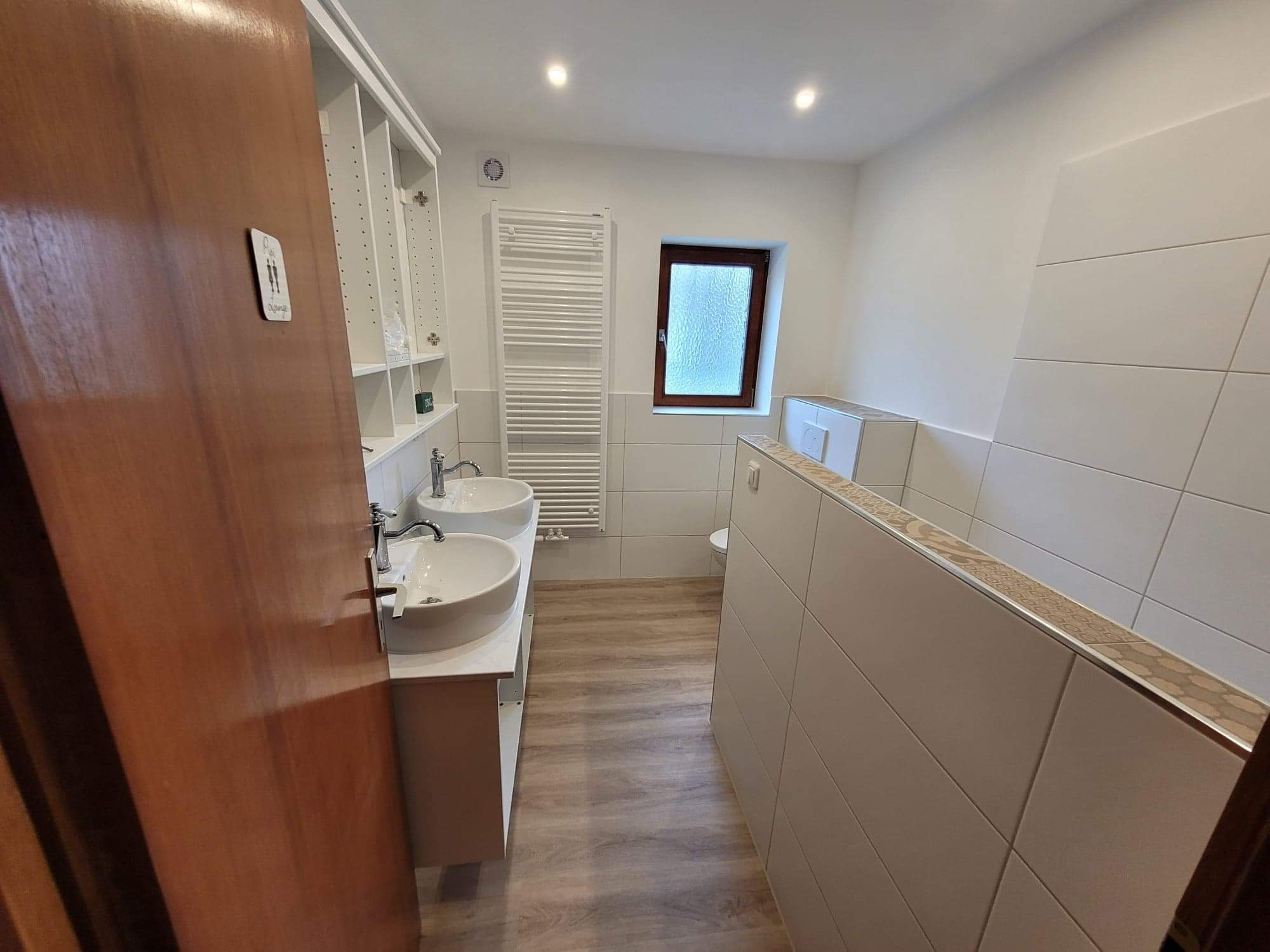 Jasna łazienka z dwoma umywalkami, grzejnikiem drabinkowym i oknem z szybą ornamentową. Na drzwiach brązowych piktogram toalety.