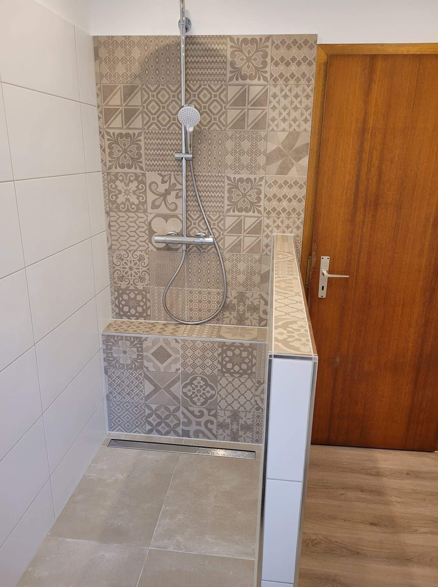 Wykończona strefa prysznicowa z deszczownicą, mozaiką z płytek w odcieniach beżu i szarości oraz odpływem liniowym, widok na drewniane drzwi.