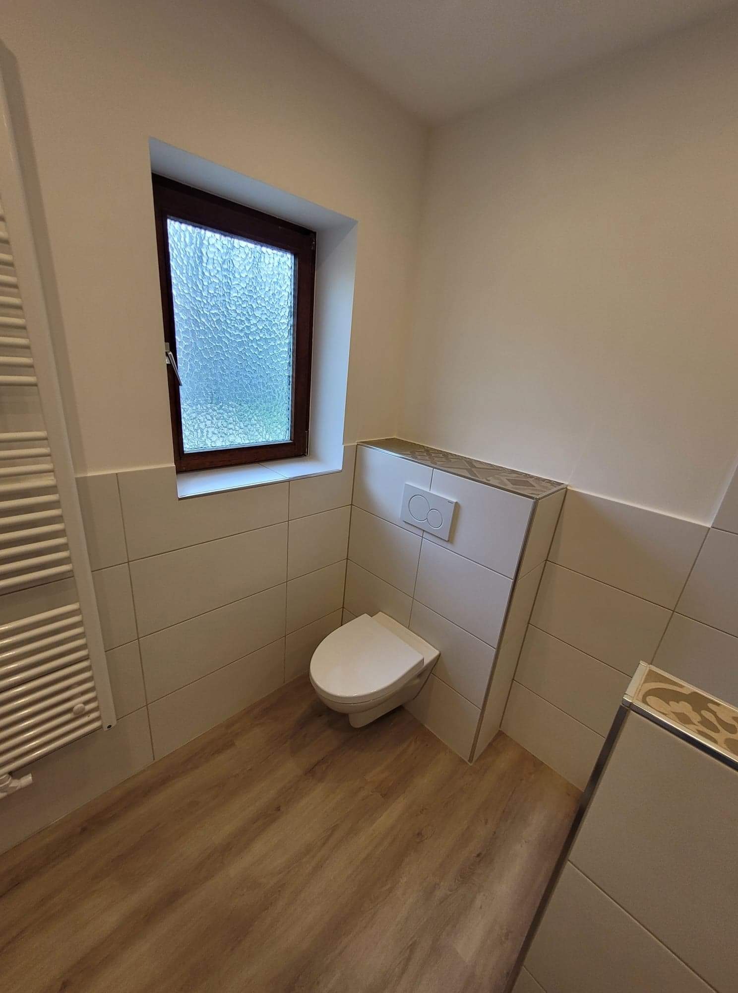 Nowoczesna łazienka po remoncie: podwieszana toaleta z przyciskiem spłukiwania, drewnopodobna podłoga, grzejnik drabinkowy, okno z teksturowaną szybą i dekoracyjne wykończenie nad zabudową toalety.