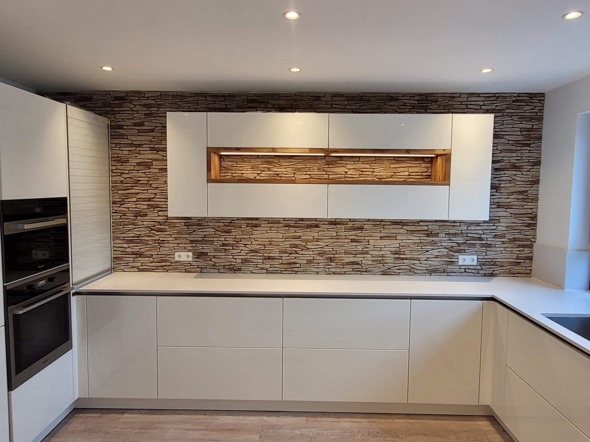 Nowoczesna kuchnia z białymi, minimalistycznymi szafkami, blatem w jasnym odcieniu i dekoracyjną ścianą z imitacji kamienia, podświetlane półki.