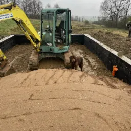 Żółta mini koparka na placu budowy z brązowym psem rasy Newfoundland stojącym obok wykopu pod fundamenty, zabezpieczonego czarną folią izolacyjną i pomarańczowymi rurami kanalizacyjnymi.