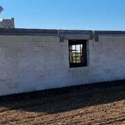 Wznoszenie ścian domu z bloczków betonu komórkowego Ytong, widoczne okna z czarną stolarką, zabezpieczone fundamenty, w tle pole uprawne i błękitne niebo.