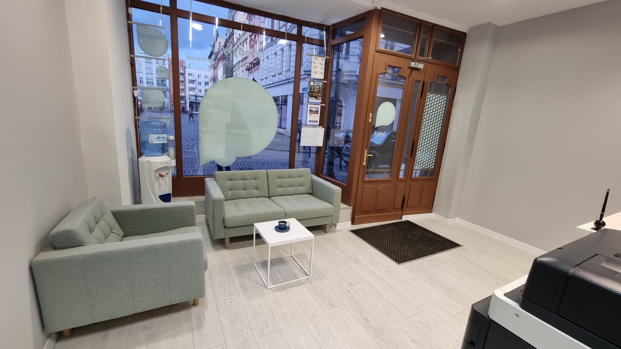 Wnętrze biura kredytowego z zielonymi sofami, stolikiem kawowym i dystrybutorem wody, widok na ulicę przez okno z naklejonymi logotypami.