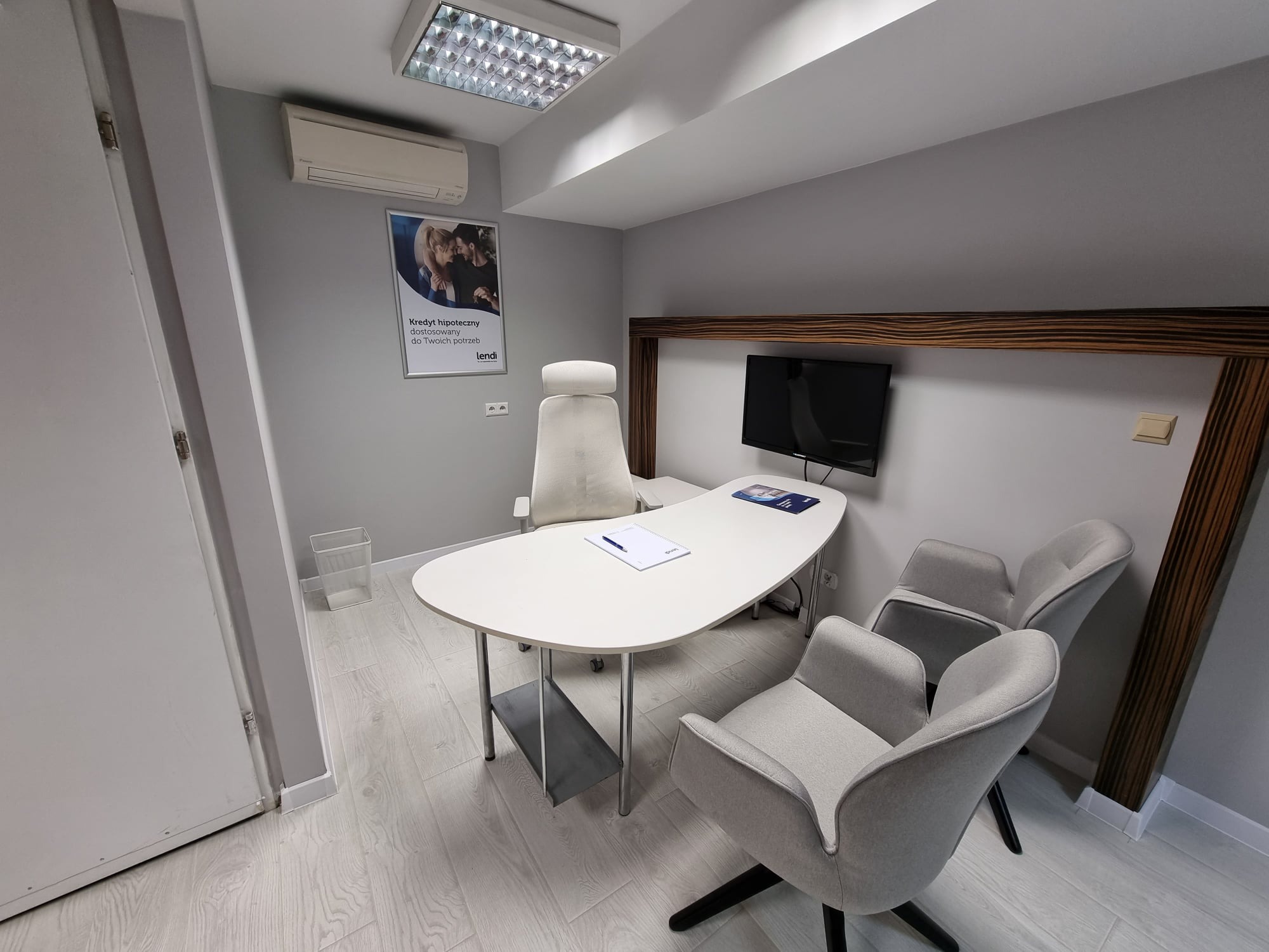 Jasne biuro z owalnym białym biurkiem, trzema szarymi fotelami dla klientów i plakatem kredytowym na ścianie, oświetlone lampą sufitową.