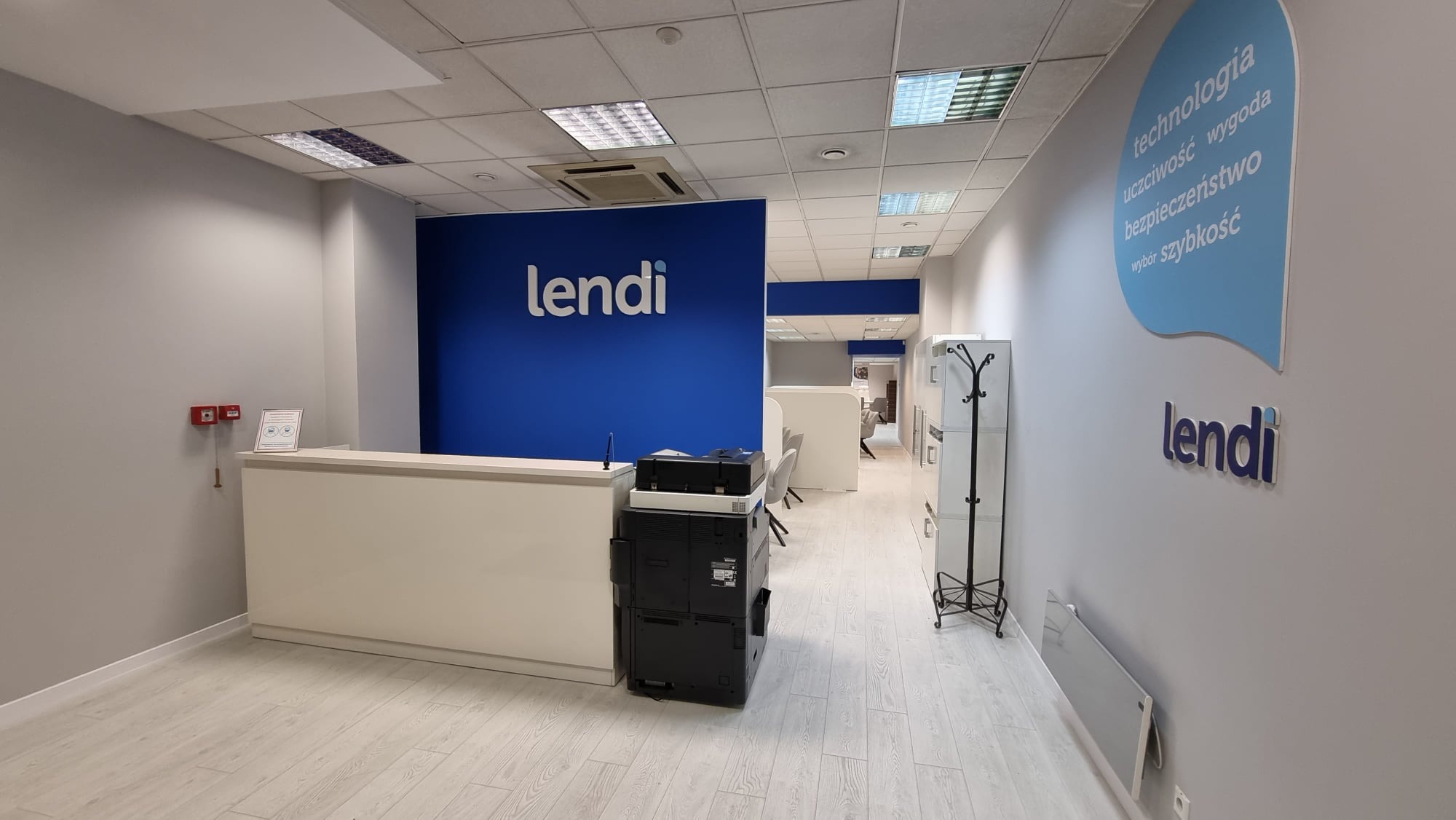 Wnętrze biura z recepcją, logo 'lendi' na niebieskiej ścianie, drukarka wielofunkcyjna obok lady recepcyjnej, hasła firmowe na ścianie, jasne podłogi.